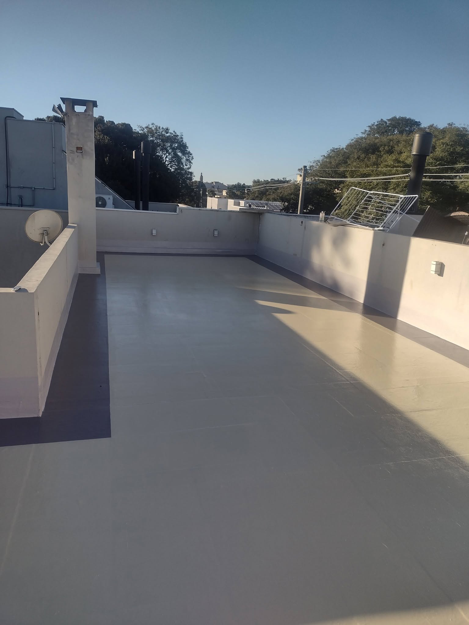 Laje de cobertura impermeabilizada com manta líquida cinza, acabamento uniforme, vista de telhados e árvores ao fundo sob céu azul, fotografia realista
