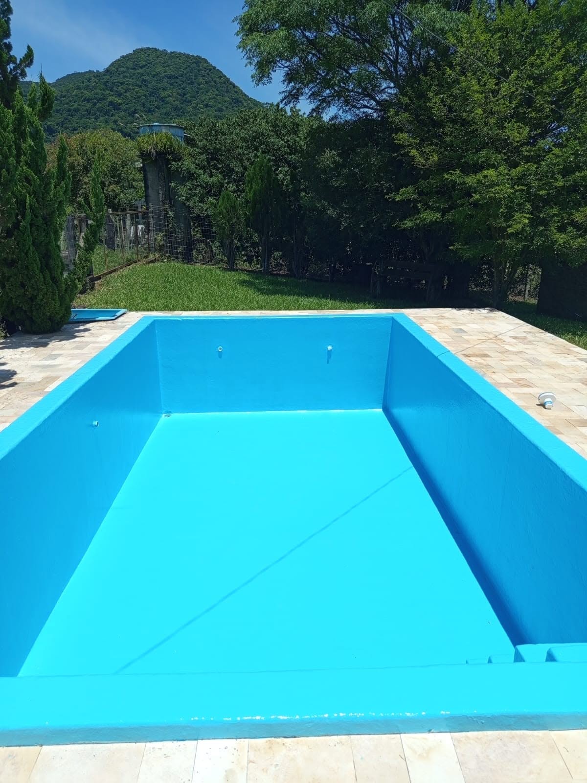 Piscina residencial impermeabilizada com manta azul vibrante, borda em pedra, jardim verde e montanha ao fundo, fotografia realista